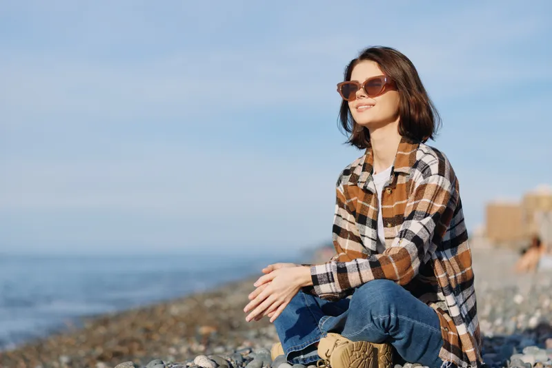 Woman wearing plaid shirt and sunglasses sits on a rocky beach by the ocean, enjoying a calm moment, casual style, sunlit scenery and blue sky above the shore.