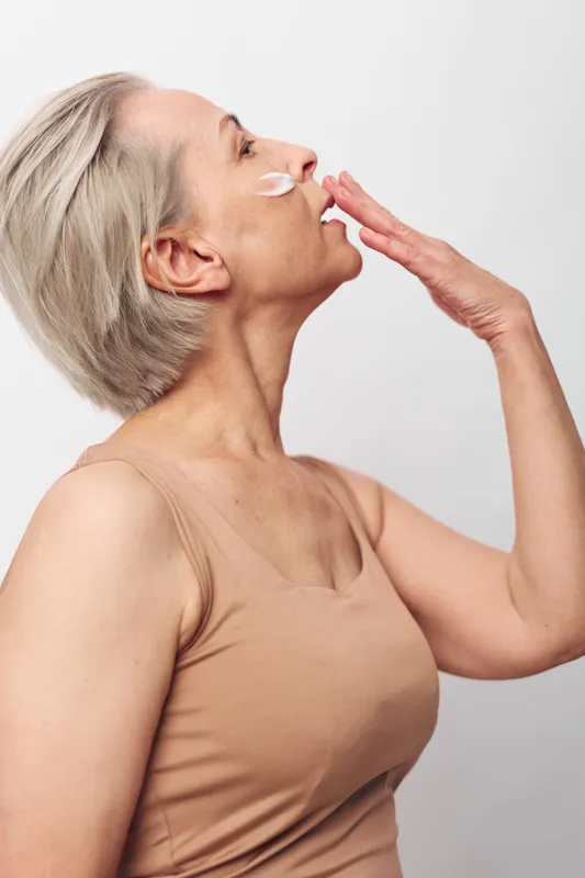 Mature woman with short grey hair applies cream on cheek, eyes closed, wearing beige tank top. Concept of skincare, aging beauty, self-care, and natural wellness in simple studio setting.