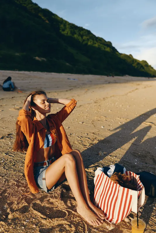 Smiling woman relaxing on sandy beach, summer vacation mood, orange jacket and casual shorts, she sits beside striped tote bag, sunlight and long shadows over sea shore, nature landscape