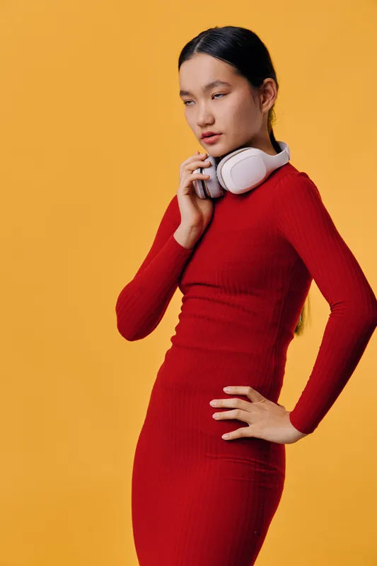 Young woman in red dress with white headphones standing against orange background looking sideways with thoughtful expression in studio. Female model with long hair and casual pose.