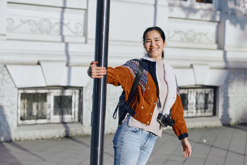Young woman leaning on a street pole with a camera bag smiling in a casual jacket and jeans urban sidewalk setting daytime light travel fashion vibe candid lifestyle shot for urban exploration