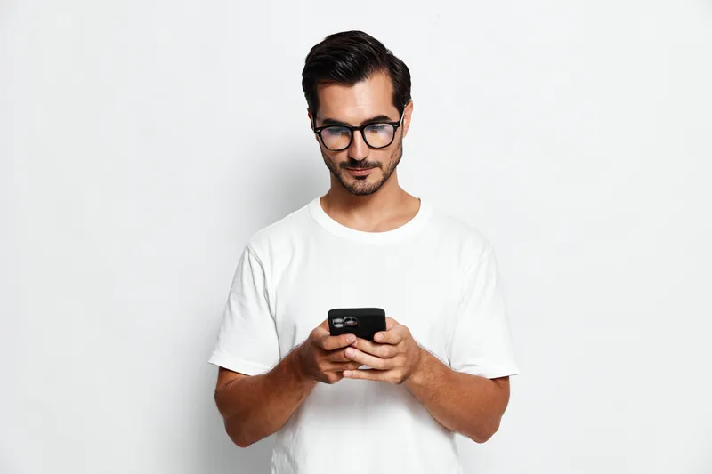 Young man wearing a casual white t-shirt focused on using a smartphone. He has glasses and stands against a plain white background, showcasing modern lifestyle.