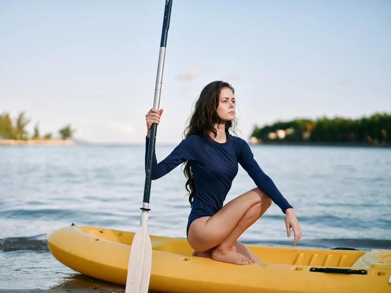 Young woman sits on a yellow kayak by the shore, holding a paddle, calm water and clear sky create a peaceful outdoor adventure moment.