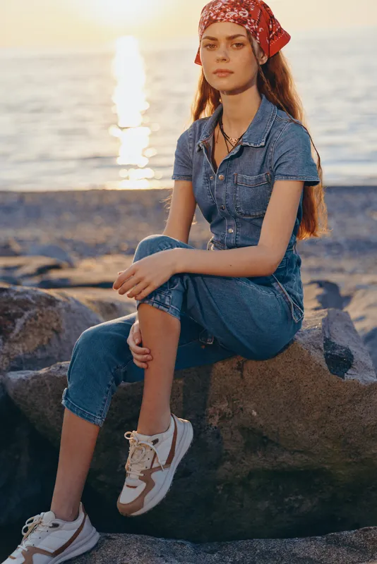 young woman sitting on rocks near sea wearing denim jumpsuit and red bandana, casual style, sunset light, relaxed mood outdoor scene