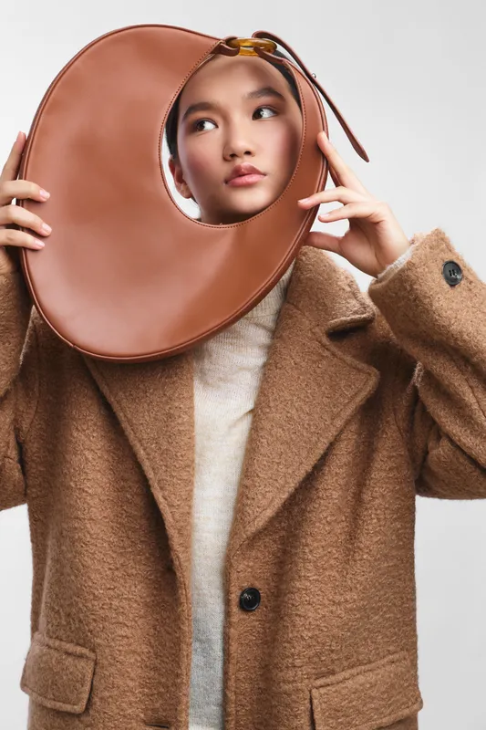 asian woman wearing a tan wool coat poses with a large circular fashion prop in a studio, creating a chic, modern mood while glancing aside with a calm, thoughtful expression.