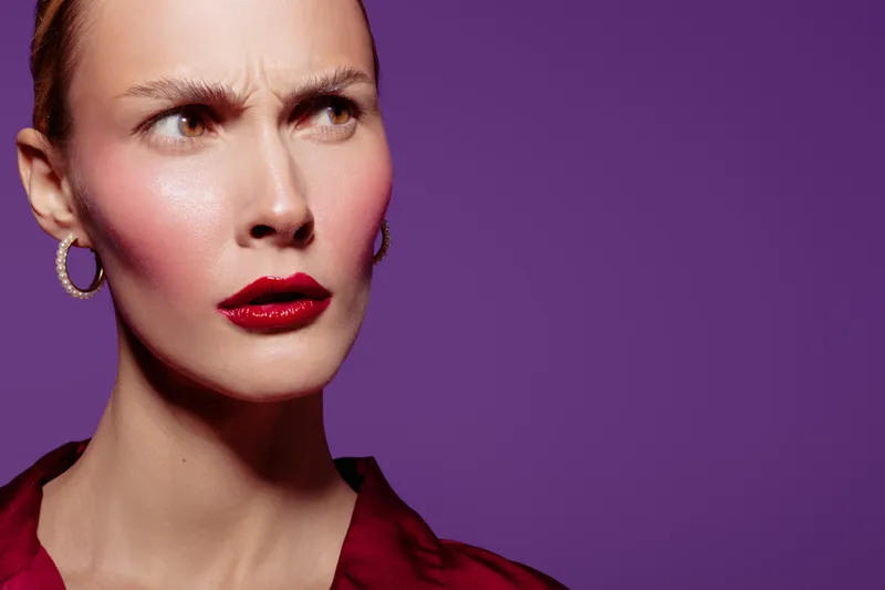 Close-up of a woman with red lipstick and hoop earrings looking confused or skeptical against a purple background. Concept of doubt, emotion, and facial expression in portrait.