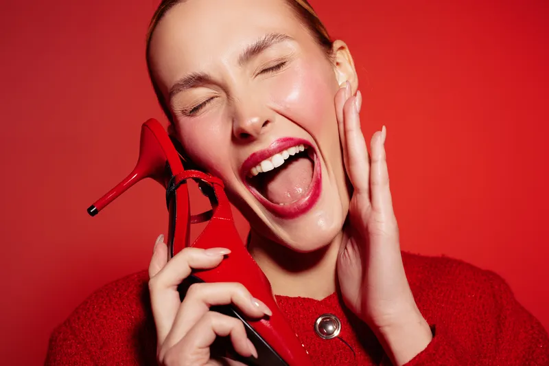 Happy woman with red lipstick and red sweater holds red high heel shoe near face, smiling with closed eyes on red background. Concept of fashion, beauty, and joyful emotions.