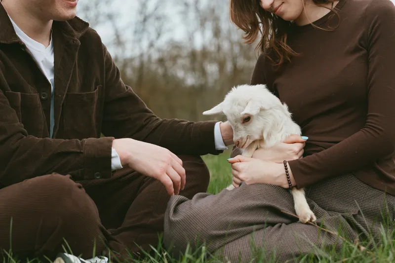 Lifestyle outdoor scene shows a couple sitting on grass, gently holding a goat kid, a calm interaction with animal and nature, warm smiles, casual clothing, peaceful family vibe and connection