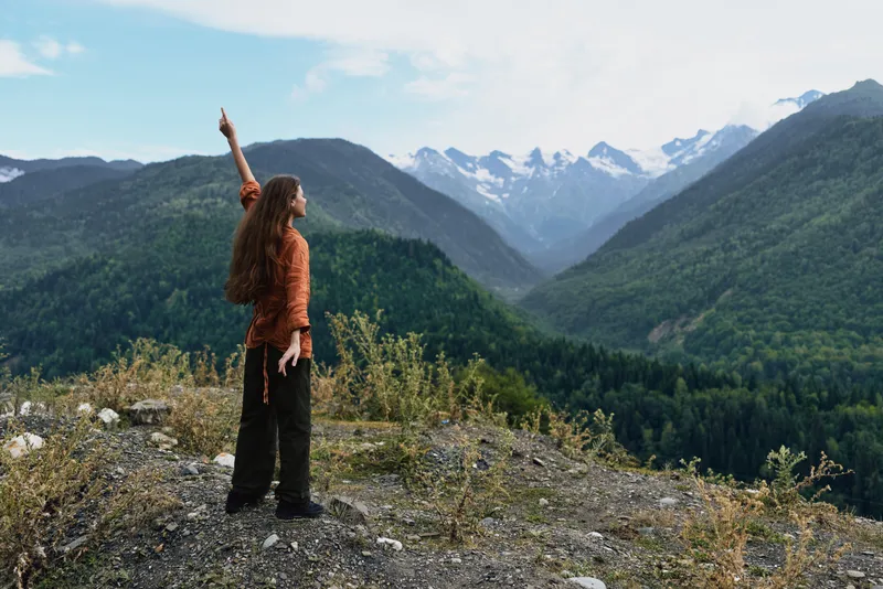 Woman stands on a rocky ridge overlooking a valley with mountains in the background, embracing outdoor adventure and nature as she points to the sky
