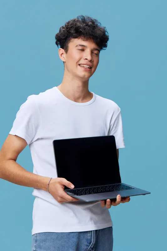Smiling young man holding open laptop with blank screen against blue background, casual clothing, curly hair, cheerful expression, technology concept.