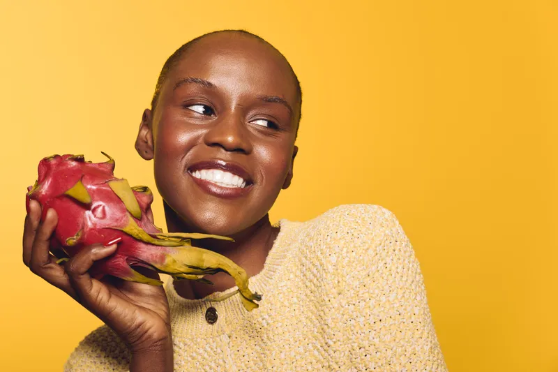 Smiling African woman with dark skin holds dragon fruit near face, wearing yellow sweater on yellow background. Concept of healthy lifestyle, fruit, nutrition, and natural beauty.