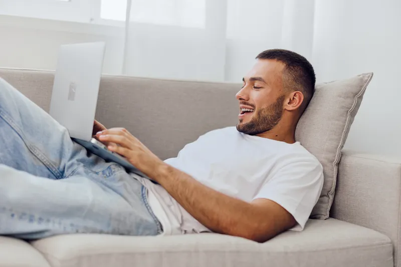 Smiling man relaxing on a couch while using a laptop, dressed in casual clothes, giving off a vibe of comfort and creativity in a bright living space.