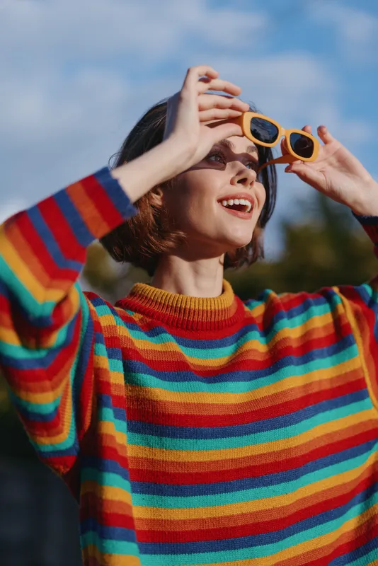 woman wearing colorful striped sweater and yellow sunglasses smiling and looking up outdoors, portrait with short bob haircut, natural sunlight and joyful casual fashion expression
