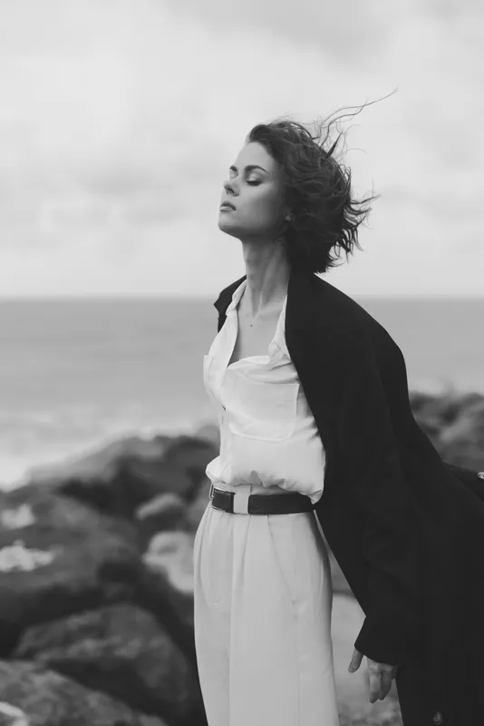 Windy coast scene with a woman in a flowing dress and cape, standing on rocks, serene coastal breeze, monochrome mood, fashion inspired