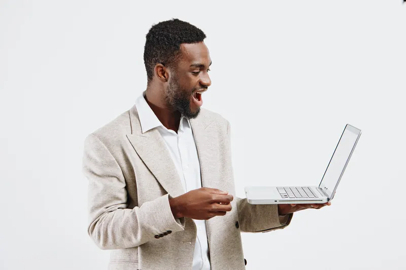 Smiling happy young African American man in business casual attire holding a laptop, looking excited and engaged, isolated on plain white background. Concept: technology, communication, success, work,