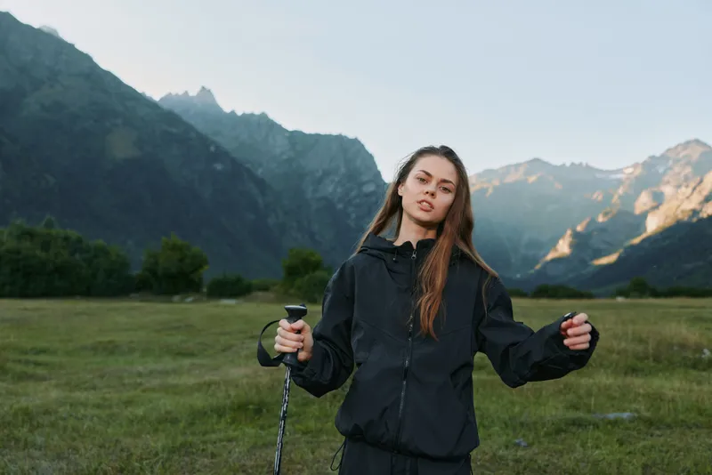 A young woman stands outdoors in a vast valley with distant mountains, holding trekking poles and wearing a dark jacket, exuding confidence and calm focus.