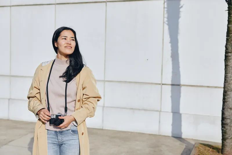 Woman with camera in an outdoor setting wearing a beige coat and denim, smiling as she photographs the surroundings, street backdrop, lifestyle photography with a candid, fashionable mood