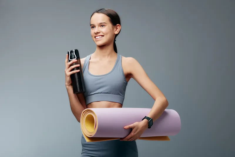 Energetic woman in athletic wear holds a water bottle and a rolled yoga mat, ready for a workout or training session. A clean studio backdrop highlights fitness, wellness and athletic determination.
