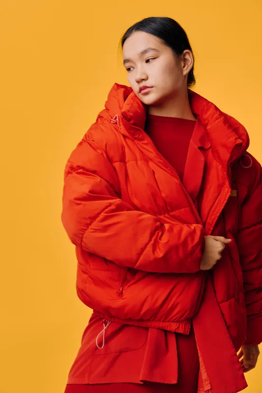 Young woman wearing a bright orange padded jacket and red sweater standing against a solid yellow background looking to the side with folded arms. Fashion, winter, casual style clothing.