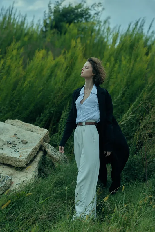 Woman walking through a grassy field in white trousers and blazer, confident pose in a natural outdoor setting, fashion and freedom, calm breeze and open space, serene countryside mood, timeless style