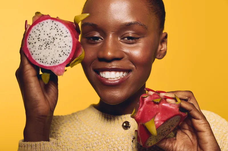 Smiling dark skinned African woman holds two halves of a dragon fruit close to her face against yellow background. Concept of healthy lifestyle, tropical fruit, and natural beauty.