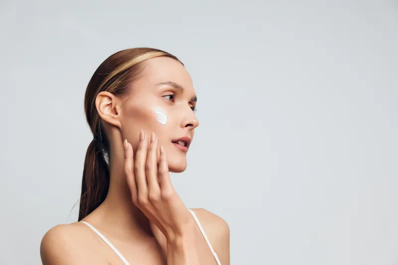 Young woman with long hair applies moisturizer cream on her cheek while looking thoughtfully to the side against a plain light background in a skincare routine moment.