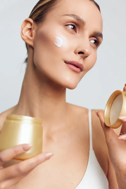 Young woman with clear skin applies moisturizing cream on her cheek while holding an open jar in a bright studio setting.
