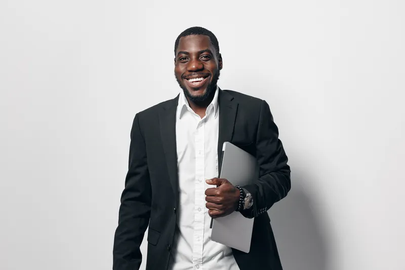 African American man with dark skin wearing black suit and white shirt holding laptop smiling confidently in studio with white background for business and technology concept.