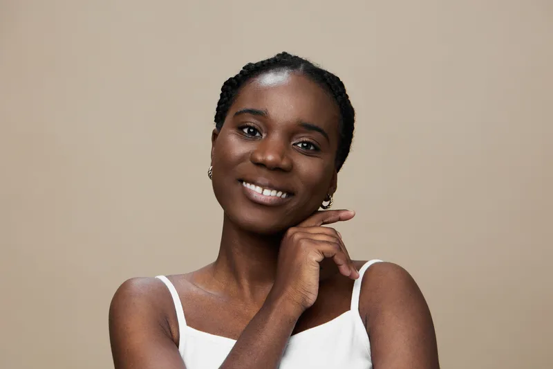 Smiling African woman with braided hair in a white tank top, showcasing confidence against a neutral beige background, perfect for beauty and fashion concepts