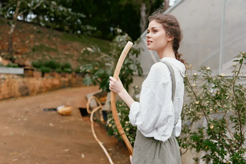 Young woman holding a wooden bow outdoors, wearing a light sweater and overalls, in a serene garden setting, reflecting nature and adventure