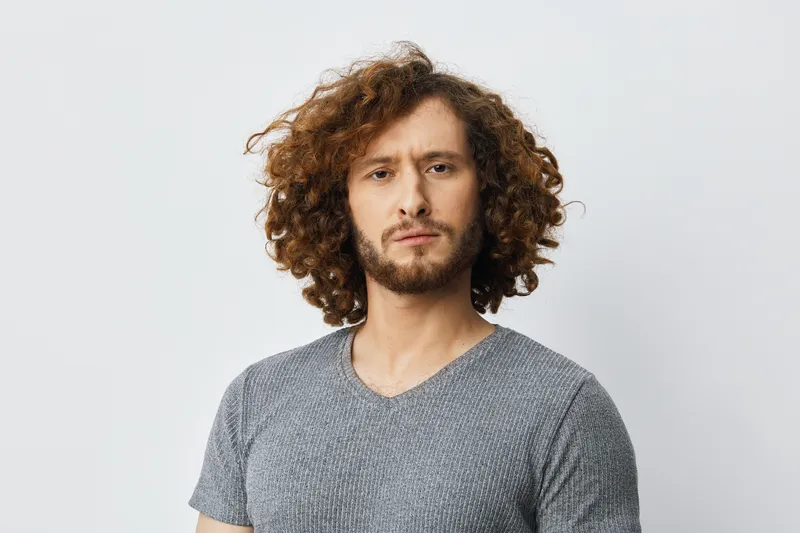 Young man with curly hair wearing a grey shirt, exhibiting a serious expression against a neutral background, ideal for concepts of personal branding and confidence