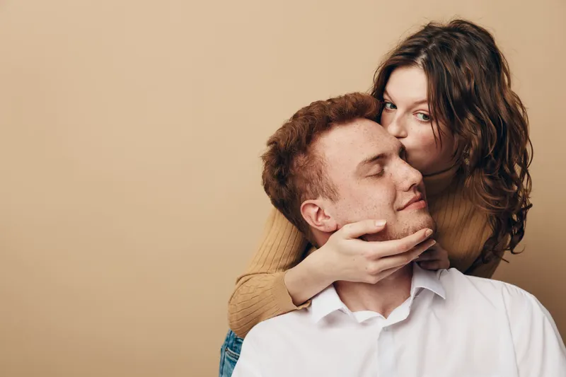 Young woman kisses man on cheek while holding his face gently. Both have relaxed expressions against beige background. Concept of love, affection, and intimate relationship.