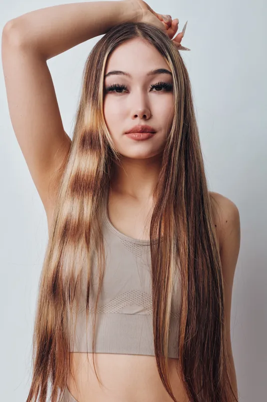 Young woman with long straight hair in beige crop top posing with hand raised on gray background. Portrait, beauty, casual style and natural makeup for fashion photography.