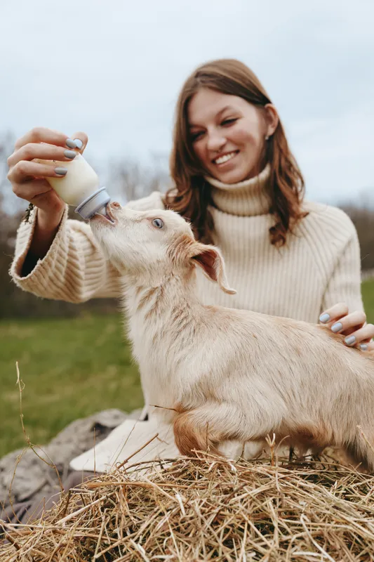 A young woman in a beige sweater happily feeds a baby goat with a bottle outdoors on a farm, capturing a tender moment of care and connection in a natural lifestyle setting.
