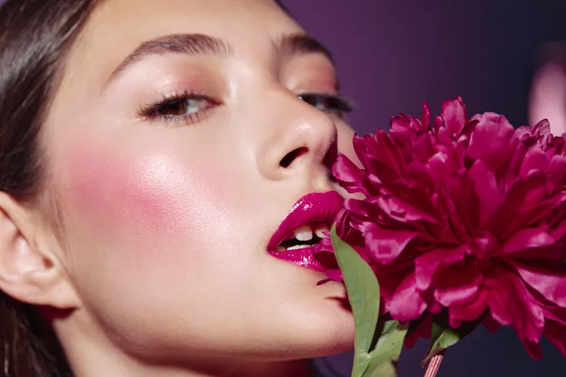Close-up of a young woman with glossy pink lips and glowing skin holding a vibrant pink flower, emphasizing beauty and fashion creativity in studio lighting on a purple backdrop.