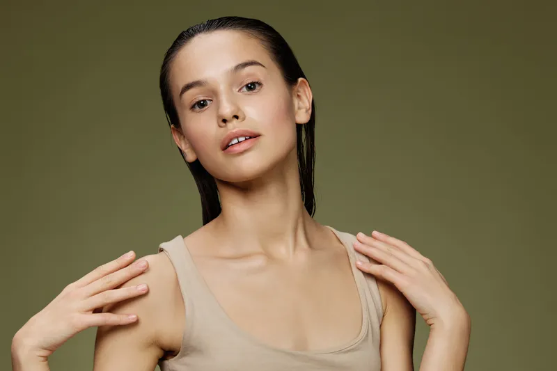 Elegant young woman posing with a natural look in soft neutral clothing, exuding confidence against a muted green backdrop, showcasing beauty and simplicity
