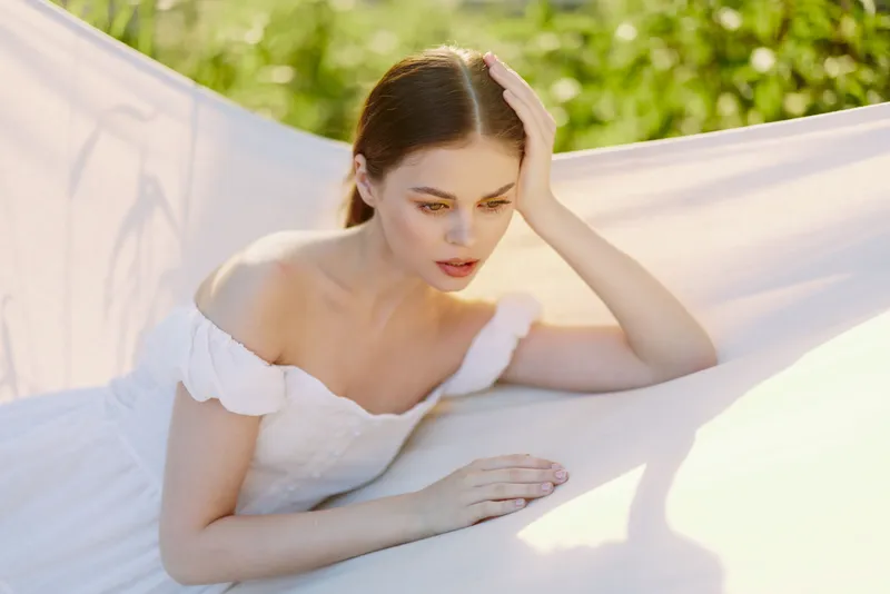Young woman in a white dress resting on a hammock with a pensive expression, surrounded by lush green nature, evoking a sense of tranquility and reflection