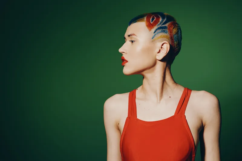 Creative woman with colorful shaved hair and red lipstick poses against a green background wearing a red sleeveless top, expressing bold and confident style.