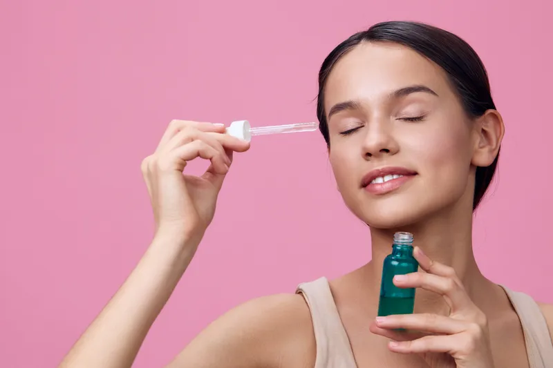 Young woman applying serum with a dropper, showcasing skincare routine with glowing skin against a pink background, representing beauty and self care