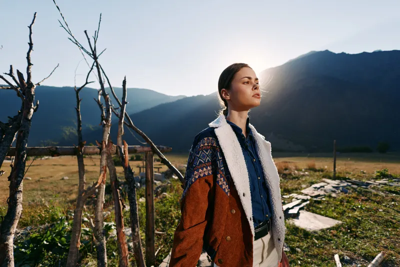 Woman portrait in mountains countryside with sunlight backlit rim wearing patterned shearling coat and denim, standing near rustic fence and meadow, fashion outdoor lifestyle with serene mood.
