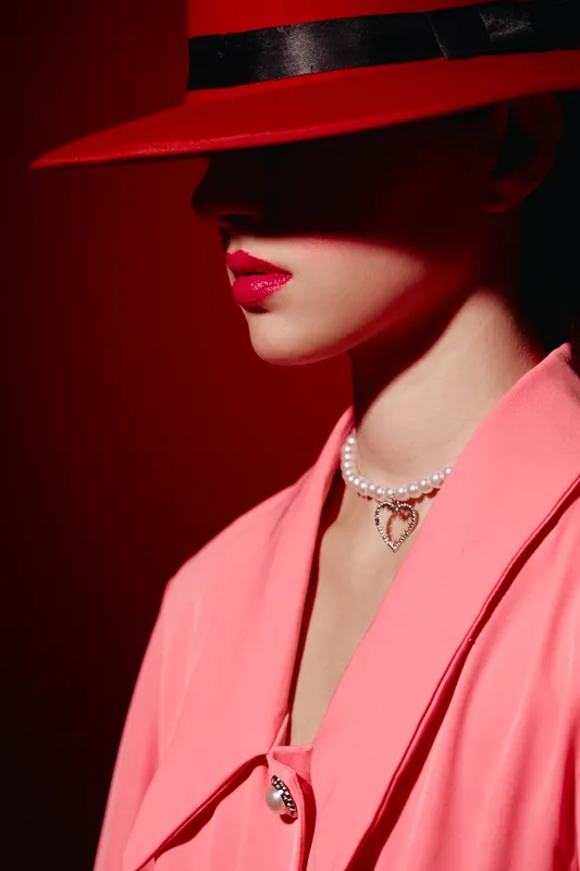 Fashion woman in red hat and bright lipstick wears pink coat and pearl necklace with heart pendant. Elegant beauty portrait with shadowed face on red background. Conceptual creative elegance.