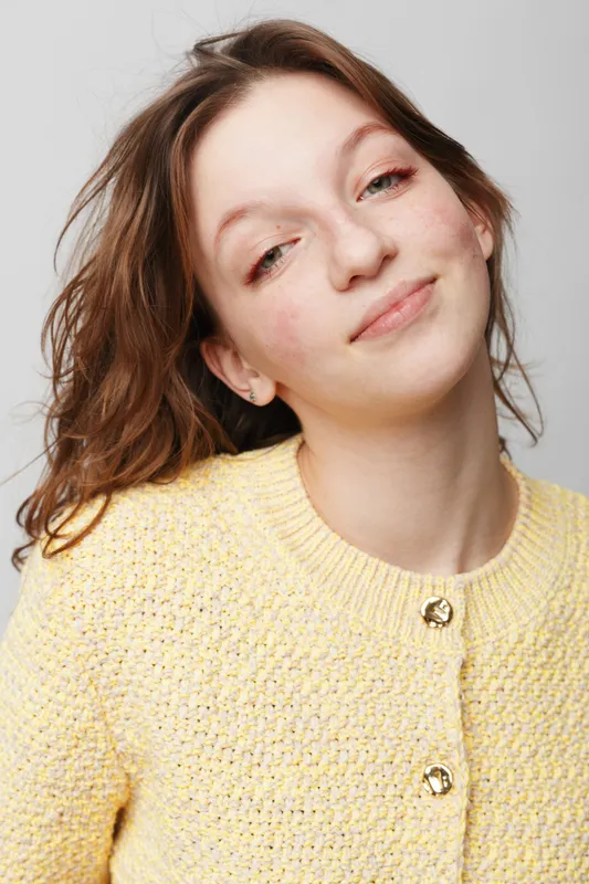 Young woman with light skin and brown hair smiles gently wearing a yellow knitted sweater. Studio portrait on white background with natural expression and casual style. Concept of happiness.