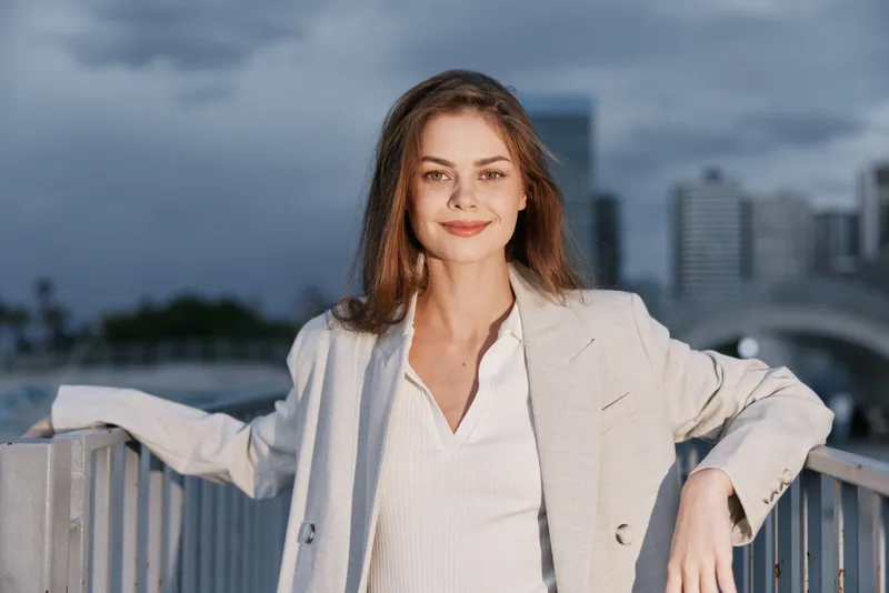 confident woman in light gray blazer posing outdoors with urban cityscape in the background during cloudy weather