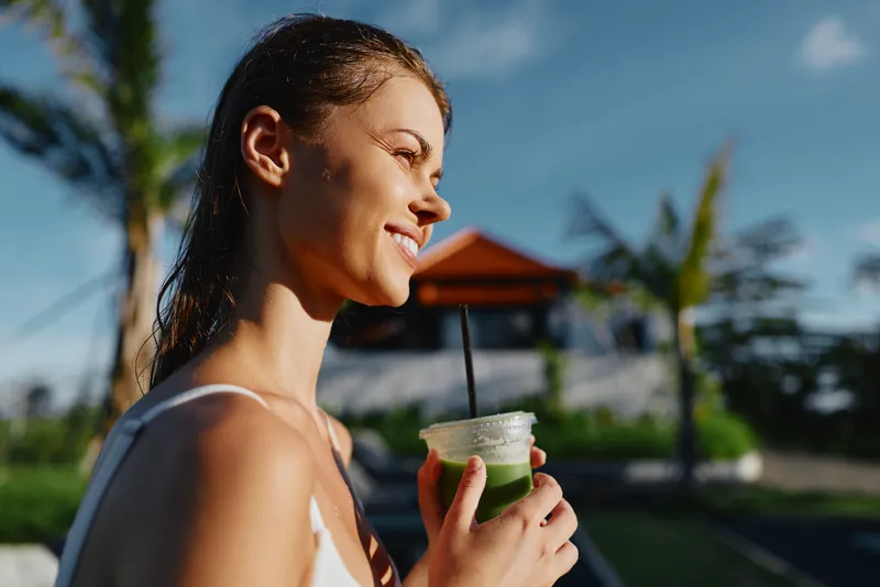 Smiling woman with wet hair holding a green smoothie outdoors on a sunny day. Tropical background with palm trees and blue sky. Concept of healthy lifestyle and wellness.