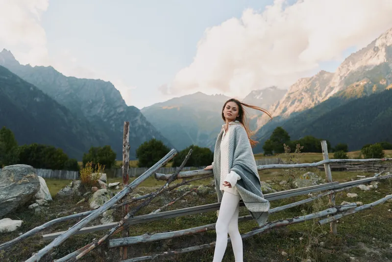 Young woman with long hair wrapped in a cozy shawl walks near a wooden fence in a mountain valley during a calm autumn day.