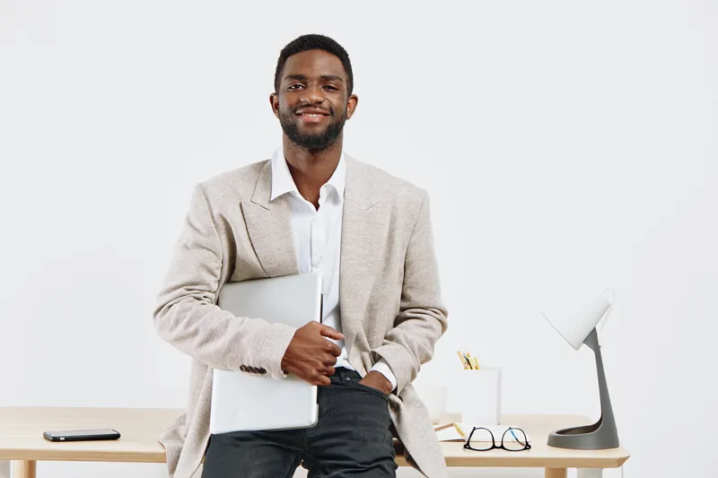 Confident young African American man in business casual attire holding laptop and smiling while standing in modern office environment. Professional, approachable, success, work, entrepreneur, lifestyl