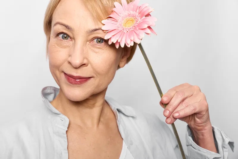 Elderly woman studio closeup holding a pink flower by her cheek, smiling gently, skincare beauty portrait on clean white background, calm spring mood, soft light, skin texture