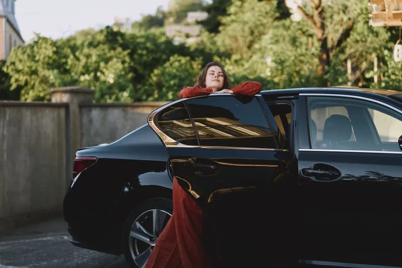 A woman wearing a red dress leans from a sleek car door, capturing a relaxed lifestyle moment outdoors with fashion, pose, and confident stance in natural light