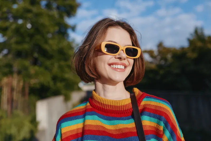 Woman in her late 20s with short hair smiling while walking outdoors in sunny park, wearing rainbow striped sweater and yellow sunglasses, casual fashion lifestyle portrait.