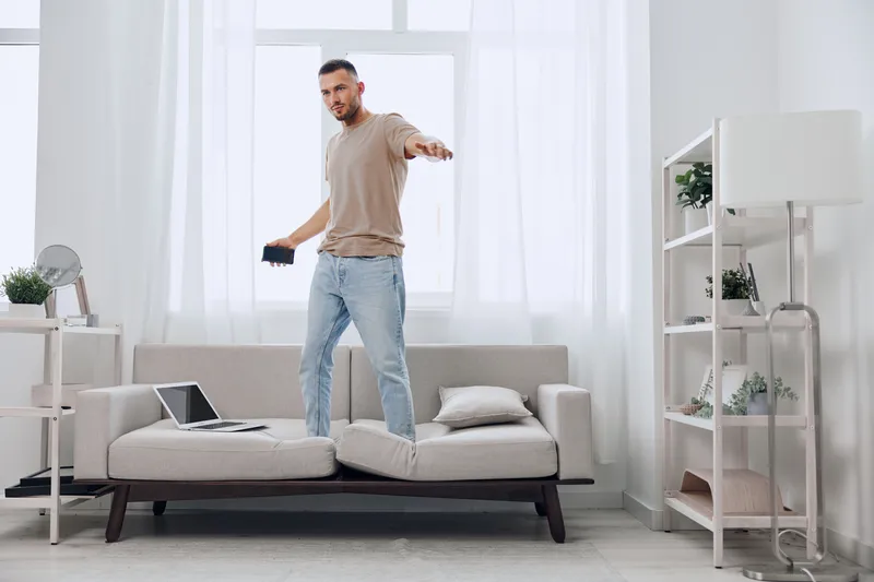 A creative man confidently expressing freedom while balancing on a couch, showcasing a modern lifestyle filled with energy and inspiration in a bright, minimalistic room.